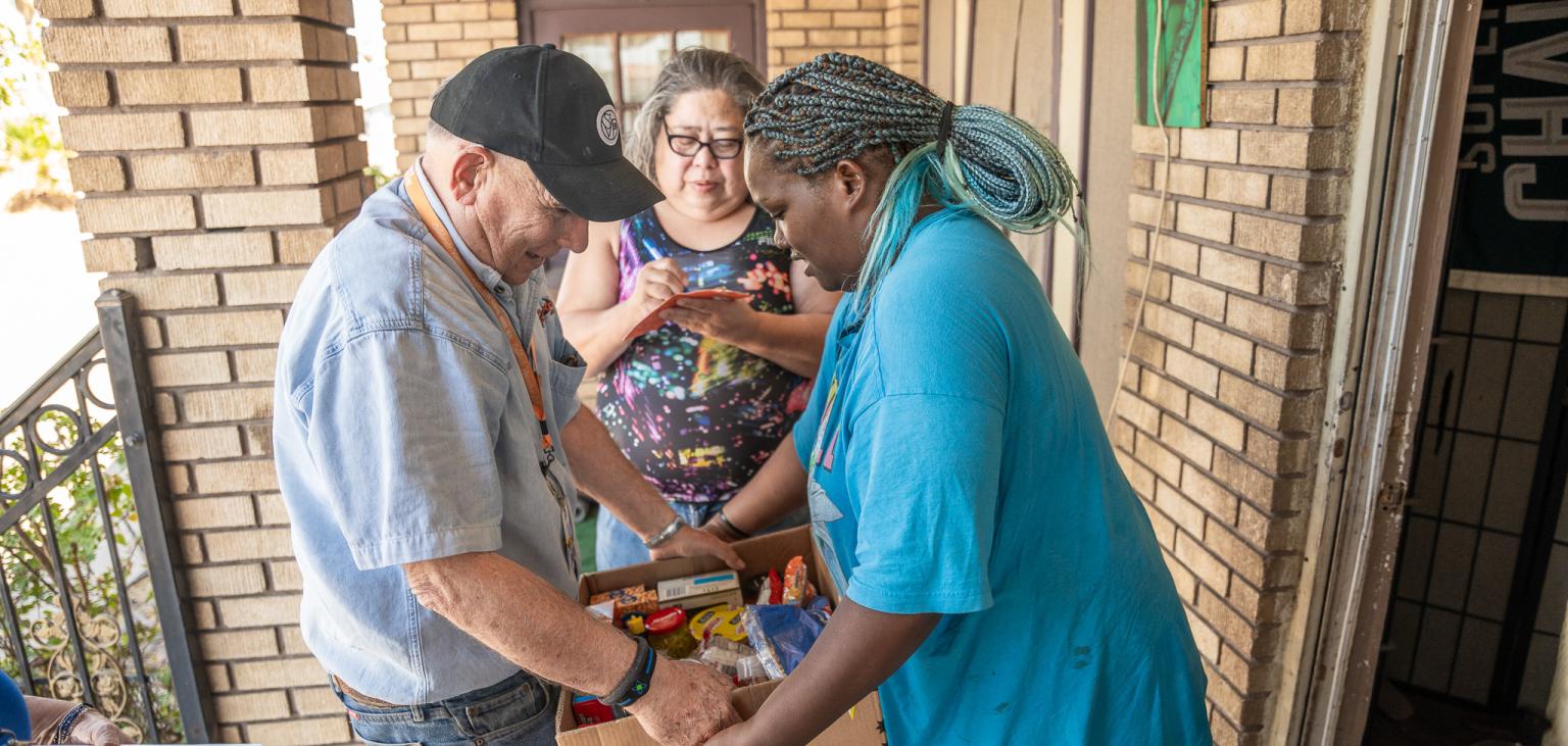 Bill, a Vincentian volunteer, hands Katherine a food box as her mother, Sandra, fills out a form in the background.