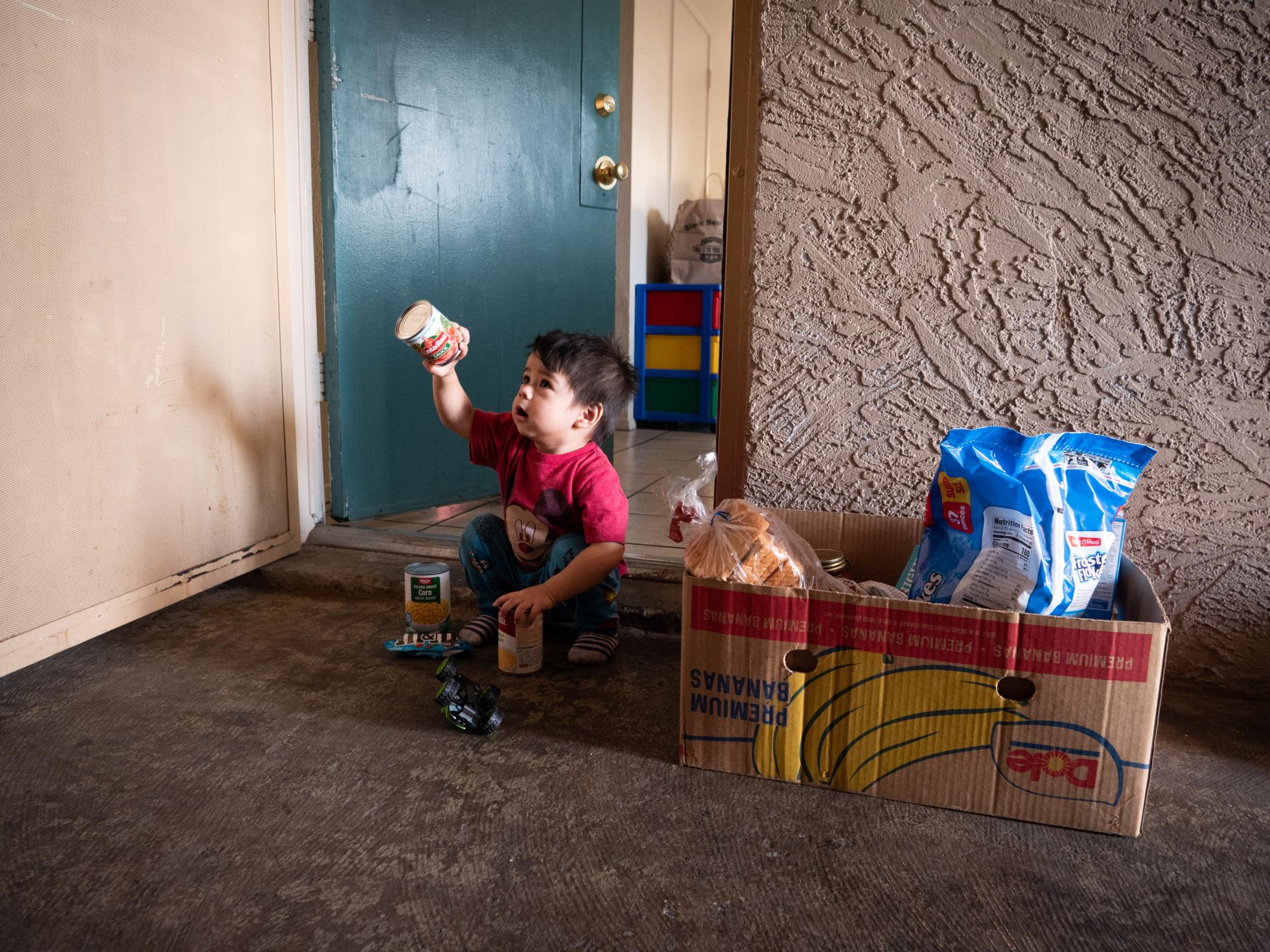 food-box-font-header Young boy sitting outside his home with a food box holding up a food item