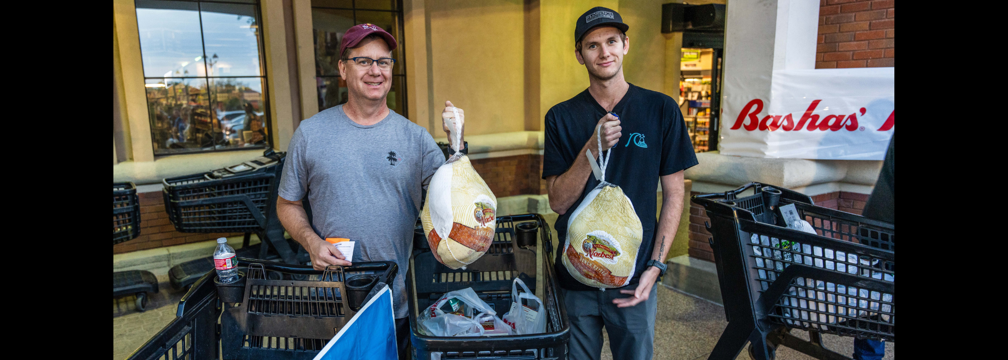Two men standing in front of a Bashas' store holding up frozen turkeys