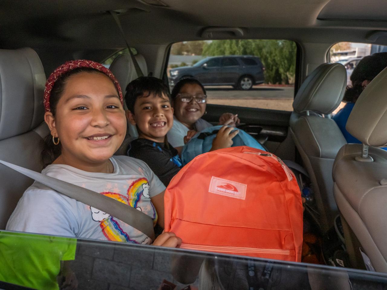 A car's backseat with three kids, each holding a new backpack given to them from SVdP.